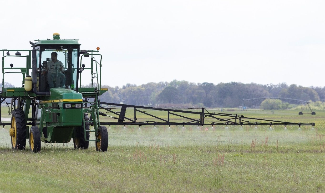 Tracker moves over grass as it sprays a liquid from many nozzles dangling at the underside of a low-hanging, metal arm-type attachment.