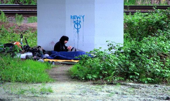 homeless youth sitting under an overpass with bedroll, bicycle.