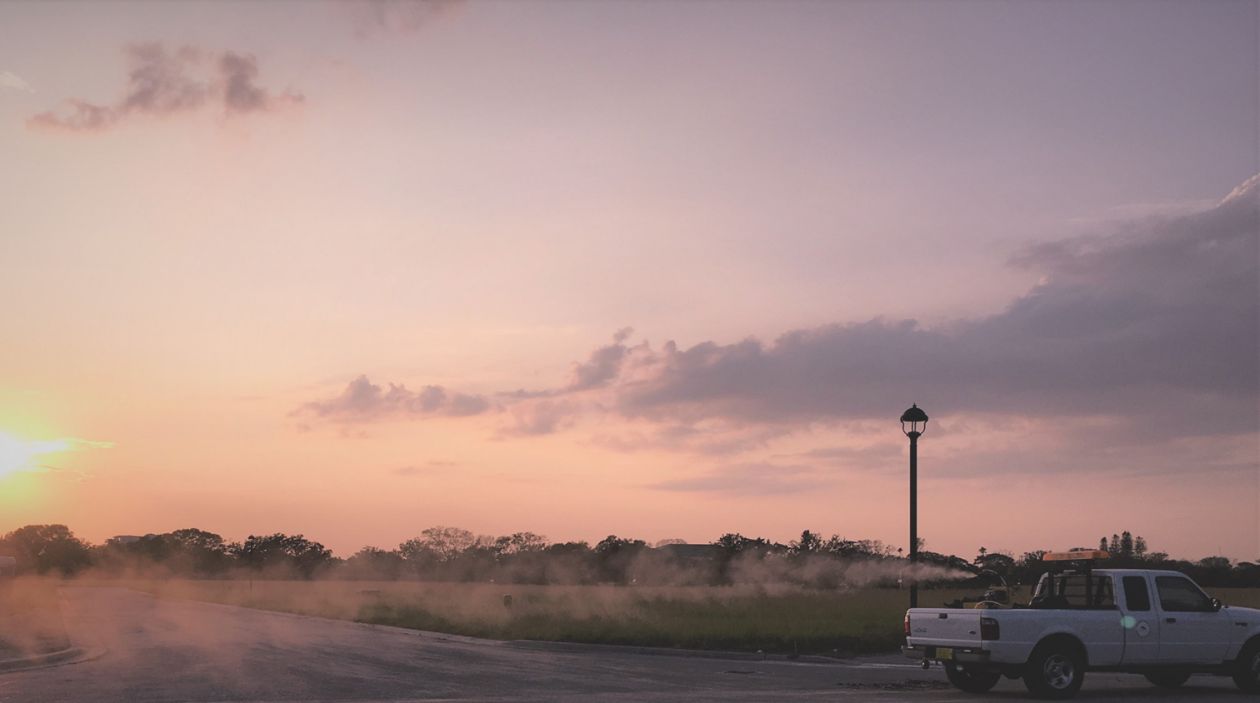 A mosquito adulticide spray cloud is released from equipment mounted in the back of a white pickup truck at sunset.