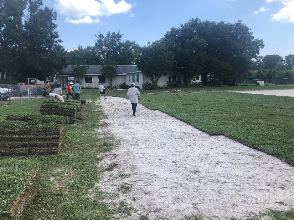 A group of people walking on the uncovered dirt between freshly placed turfgrass and stacks of the grass that will be laid down over that dirt.