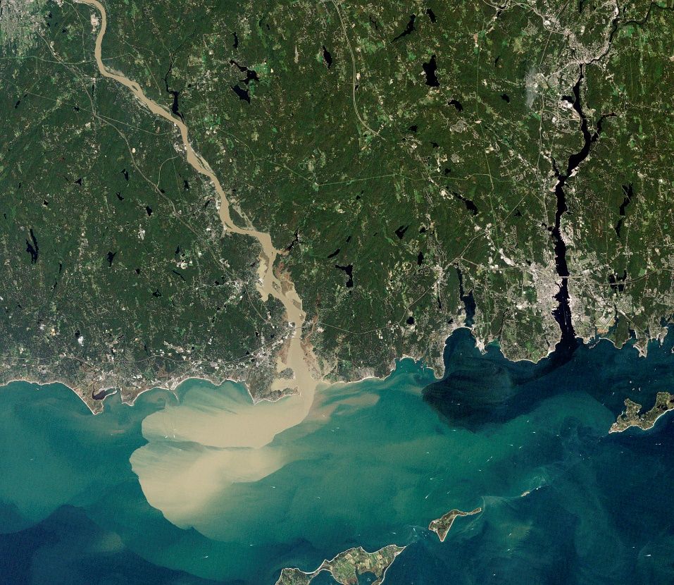 Aerial view of two rivers emptying into a larger water body: one (left) ejects water with a sand-colored cloud into the larger water body, while the other river (right) spits out a slightly darker color of water than the bigger water body.