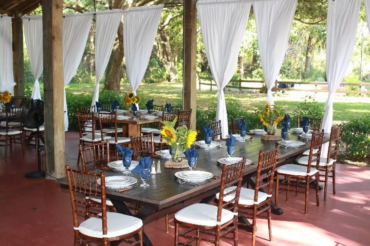 Figure 2. Nonresidential farm building prepped for dinner. The interior of a nonresidential farm building with no walls. White tie-back curtains are hung where a wall might be. Inside the building are long rectangular wooden tables with cane-back chairs with white seat pads. White place settings and blue napkins.